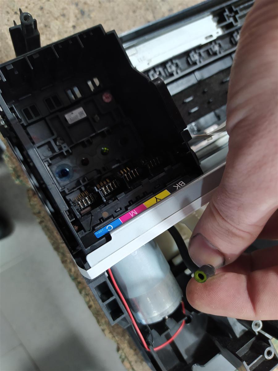 Carro de la Epson abierto mostrando los cuatro cartuchos CMYK (cian, magenta, amarillo, negro). La mano sostiene el tubo negro de salida de la capping station con el fitting verde en el extremo — ese es el tubo del que hay que aspirar con la jeringa
