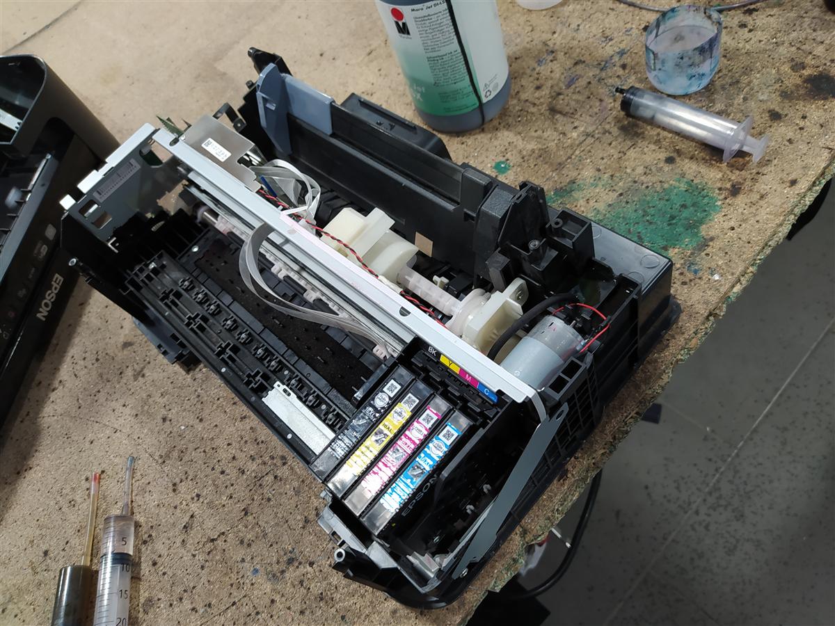Impresora Epson completamente abierta en la mesa del taller durante el proceso: carcasa quitada, cartuchos instalados, jeringa grande apoyada en la mesa y restos de tinta cian en el banco de trabajo