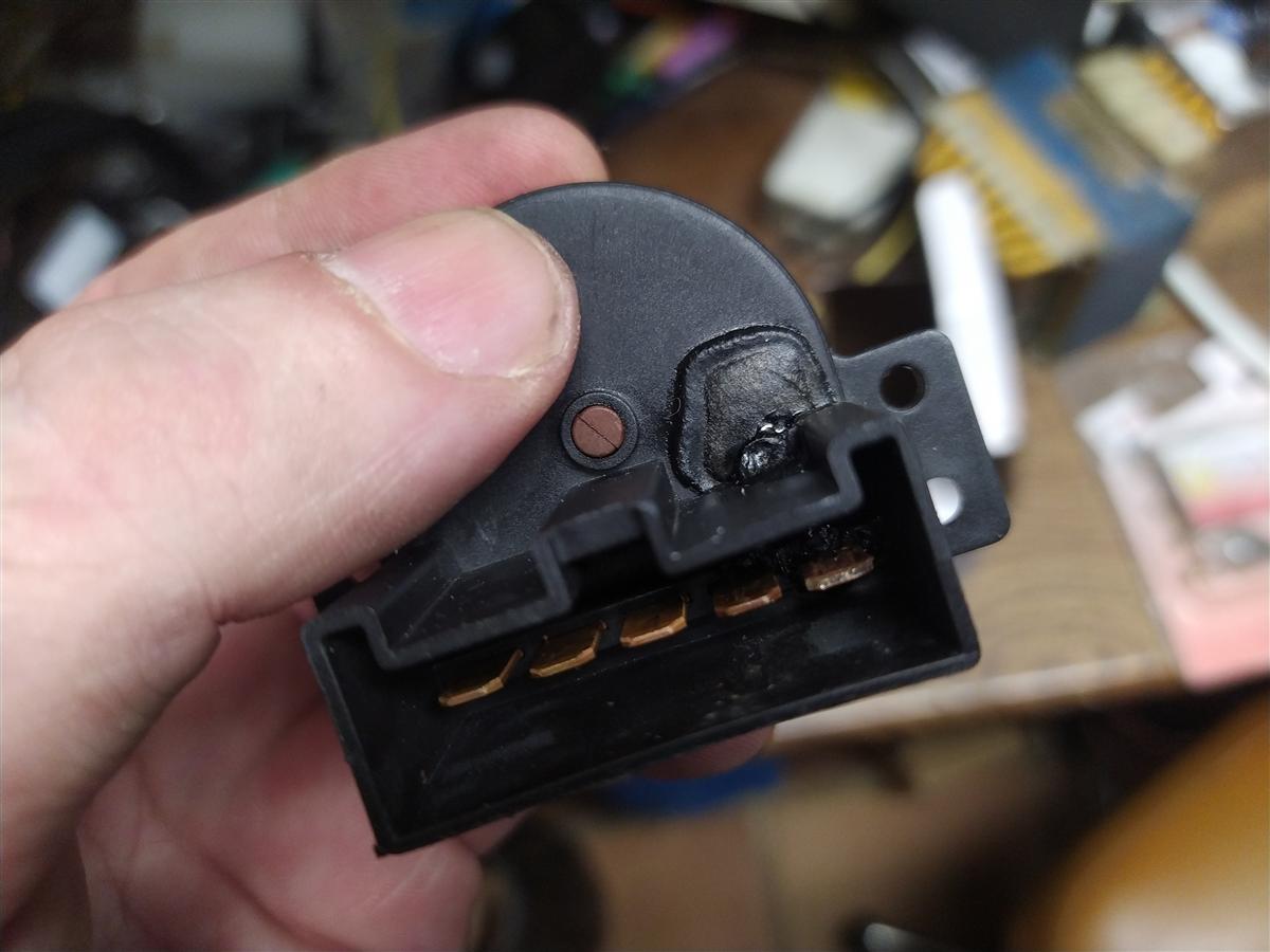 Selector desmontado sostenido en mano, mostrando la quemadura negra en la pista de carbono junto al pasador central y los cuatro contactos dorados del conector en la parte inferior